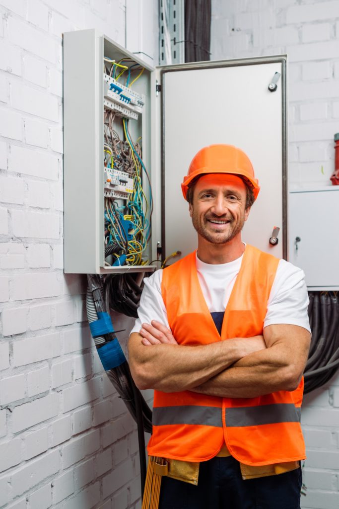 Handsome electrician in hardhat and safety vest smiling at camera near electrical distribution box